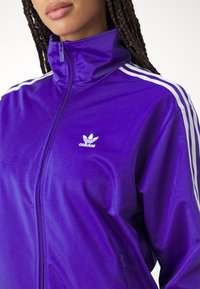 Lila zip-up trackjacka i ett slätt material, med vita ränder på axlarna och en liten vit Adidas-logga på bröstet.