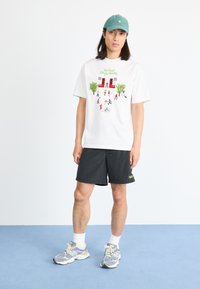 Hvit bomulls T-skjorte med fargerik grafikk som viser figurer og trær; brukt med sorte shorts og lyse treningssko.