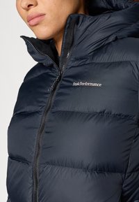 Tumed sinine täispuhutav jakk horisontaalsete tikanditega, kõrge kaelusega ja täispika tõmbelukuga. Rinnal on heledast niidist Peak Performance'i logo.