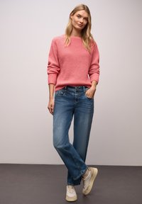 Pull rose tricoté avec un col rond, associé à un jean en denim bleu clair et des baskets beige, debout devant un fond neutre.