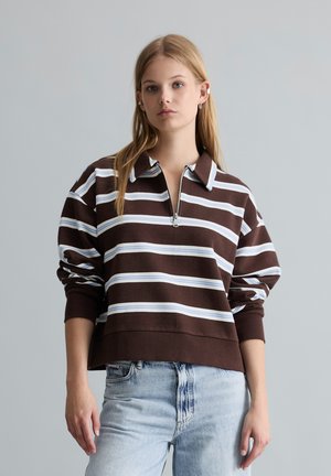 Jeune femme portant un pull zippé rayé marron et blanc et un jean bleu clair, debout devant un fond gris uni.