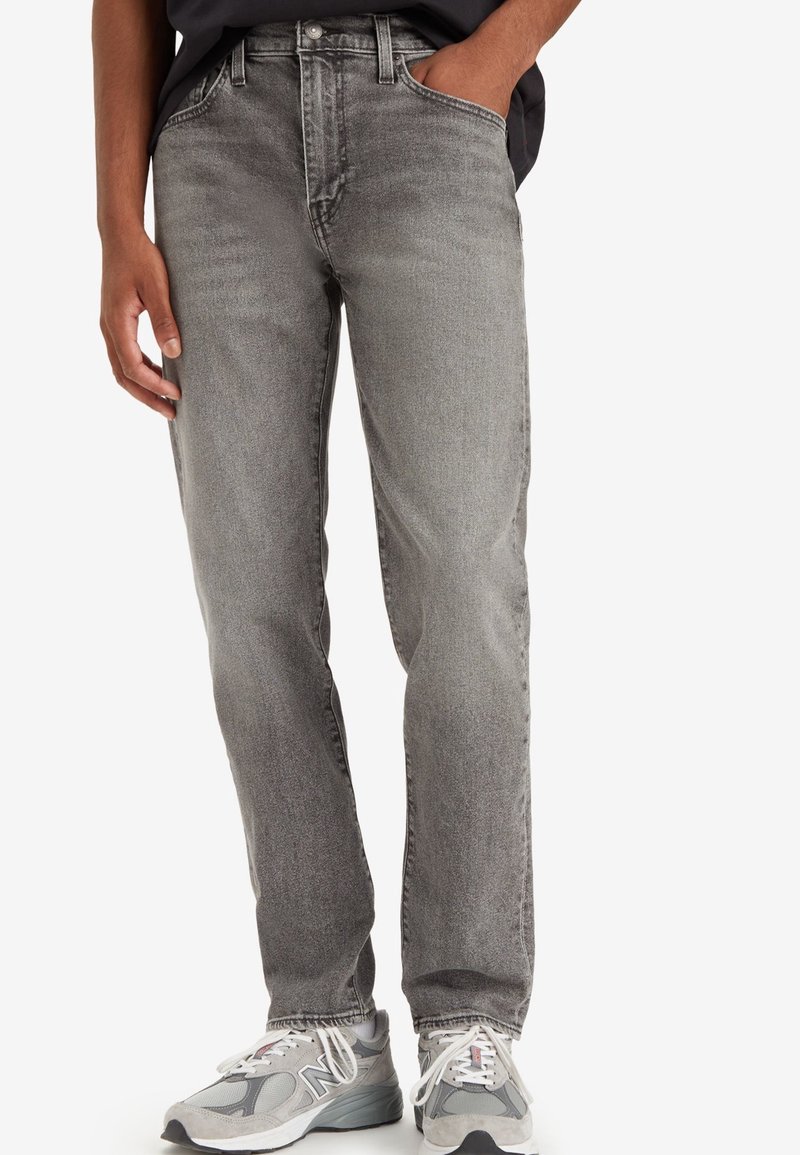 Homme portant un jean gris délavé à jambes droites, une chemise noire, une main dans la poche, et des baskets grises New Balance.