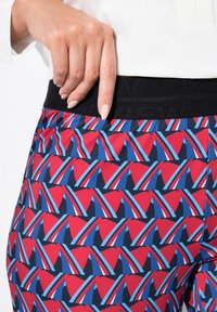 Pantalones con estampado rojo y azul, con una cinturilla elástica negra, que presentan un diseño geométrico de triángulos y rayas, hechos de una tela suave.