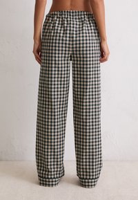 Persoon met hoge taille, losse zwarte en witte geblokte broek, gezien van achteren tegen een eenvoudige muur achtergrond.