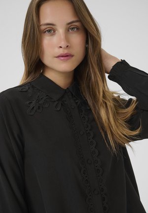 Giovane donna con lunghi capelli castano chiaro indossa una blusa nera con colletto in pizzo floreale e una placca di bottoni decorativa, con una mano che tocca i capelli.