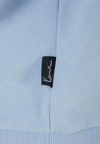 Lyseblå strikket stof med ribbet detalje ved kanten. Sort label med hvid kursiv tekst syet ind i sidesømmen. Struktureret overflade.