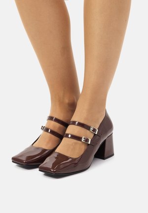 Des escarpins en cuir verni marron avec des orteils carrés, deux brides réglables et un talon épais. Texture lisse avec une finition brillante.