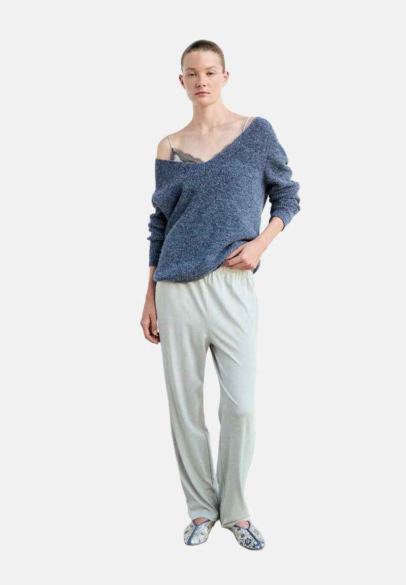 Femme portant un pantalon gris ample, un pull bleu à épaules dénudées dévoilant un caraco en dentelle noire, et des pantoufles à motifs, debout avec une main dans la poche.