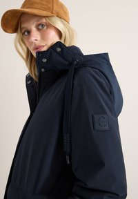 Giacca con cappuccio blu navy, dalla consistenza liscia, con coulisse regolabile, chiusure a scatto e un'etichetta nera con logo sulla manica.