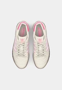 Adidas teniske z kremnim zgornjim delom, roza črtami, perforiranim prstnim delom in sivo gumijasto podplatom. Šnure so svetlo sive z roza poudarki.