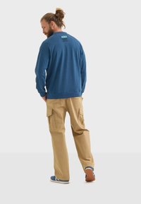 Felpa blu a girocollo con etichetta sul retro, abbinata a pantaloni cargo color sabbia con tasche laterali e vestibilità comoda, indossata con scarpe blu.