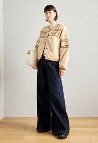 Beżowa bluza z polaru z kolorowymi poziomymi paskami, dużymi guzikami i bocznymi kieszeniami, połączona z szerokimi ciemnymi dżinsami i czarnymi butami.