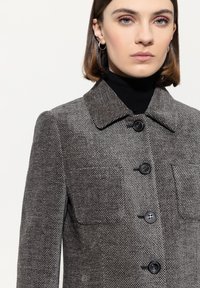 Frau mit kurzen braunen Haaren, die einen grauen strukturierten Mantel mit schwarzen Knöpfen und einen schwarzen Rollkragenpullover vor einem hellen Hintergrund trägt.