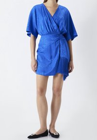 Blue wrap dress with short kimono sleeves, textured fabric, gathered waist, and fringed hem detail; paired with black flats.