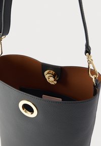 Μαύρη δερμάτινη τσάντα τύπου κουβά με χρυσή κυκλική λεπτομέρεια και μαγνητικό κλείσιμο. Εσωτερική επένδυση σε καφέ χρώμα, με ετικέτα μάρκας. Ρυθμιζόμενο λουράκι.