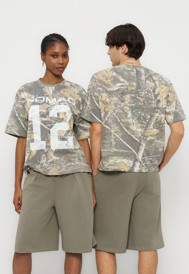 Δύο μοντέλα φορούν ταιριαστά oversized t-shirts με καμουφλάζ, ένα κοιτάει προς τα εμπρός με το κείμενο "NOMAD 12" και χαλαρά χακί πράσινα σορτς.