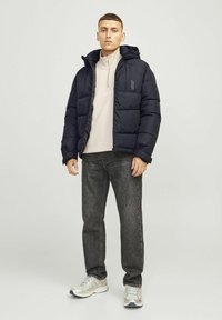 Sort puffet jakke med hætte, let materiale, lynlås lukning og sidelommer, båret over en beige zip-up top og grå jeans.