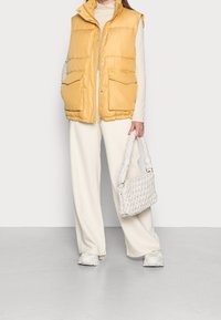 Gelbe Pufferweste mit hohem Kragen und Fronttaschen, getragen über einem cremefarbenen Pullover. Kombiniert mit weißen weiten Hosen und einer geflochtenen Handtasche.