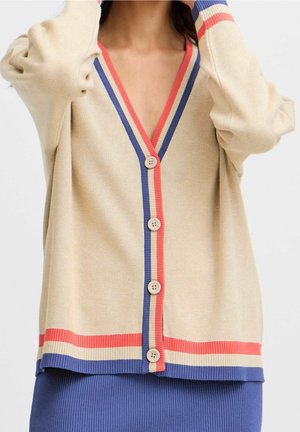 Beżowy kardigan z niebiesko-czerwonymi paskami przy krawędziach, zapinany z przodu na guziki, noszony na niebieskiej prążkowanej spódnicy.