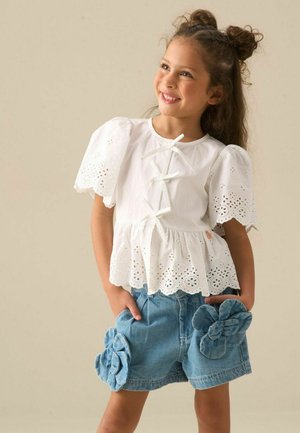 Jeune fille souriante aux cheveux bruns coiffés en chignons porte un chemisier blanc à œillets avec des manches bouffantes et un short bleu décoré de grandes fleurs en tissu.
