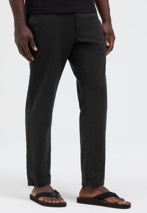 Mann trägt schwarze gerade geschnittene Hose und schwarze Flip-Flops, steht vor schlichtem hellem Hintergrund.