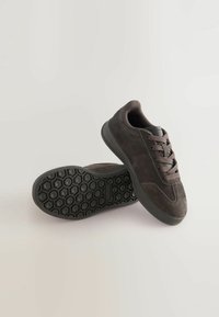 Para ciemnoszarych zamszowych butów sportowych z fakturowanymi czarnymi gumowymi podeszwami i dopasowanymi szarymi sznurówkami na neutralnym tle.