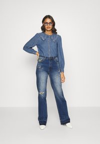 Camisa de ganga com colarinho franzido, botões na frente. Jeans de cintura alta azul claro com detalhes desgastados e bordados coloridos nos bolsos.