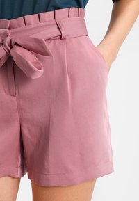 Pantaloni rosa su misura con vita alta, cinturino in tessuto, tasche laterali e una texture liscia. L'orlo è dritto e arriva a metà coscia.
