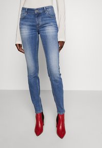 Vaqueros skinny de denim azul con un acabado desgastado, cinco bolsillos y cierre de botón, combinados con botas de tobillo rojas con tacón.