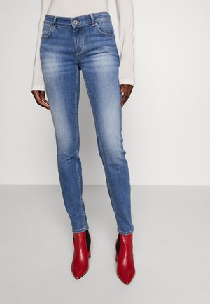 Vaqueros skinny de denim azul con un acabado desgastado, cinco bolsillos y cierre de botón, combinados con botas de tobillo rojas con tacón.