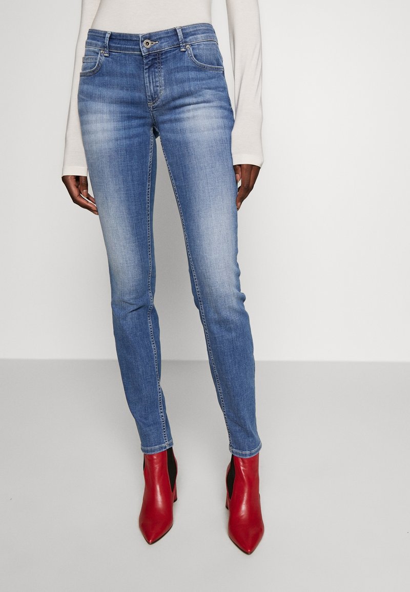 Vaqueros skinny de denim azul con un acabado desgastado, cinco bolsillos y cierre de botón, combinados con botas de tobillo rojas con tacón.