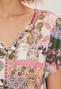 Bluzka wielorzędowa z dekoltem w kształcie litery V, z kwiatowymi i geometrycznymi wzorami w odcieniach różu, zieleni i kremu, o miękkiej teksturze materiału.