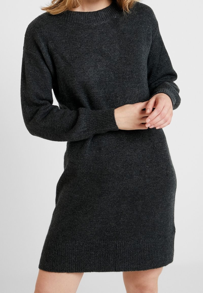 Femme portant une robe pull à manches longues gris foncé avec des poignets et un ourlet côtelés, les mains doucement croisées à la taille.