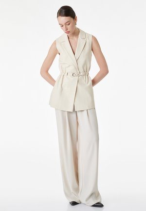 Frau trägt ärmellosen cremefarbenen Blazer mit Gürtel und weit geschnittene cremefarbene Hose sowie schwarze spitze Schuhe, steht mit den Händen hinter dem Rücken.