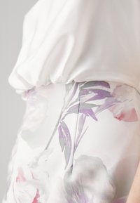 Blouse en satin blanc avec une taille froncée, associée à une jupe à motif floral dans des teintes douces de rose et de violet. Texture lisse et design délicat.