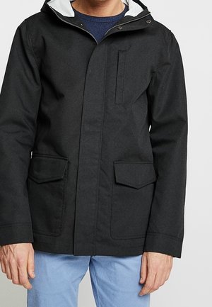 Mand iført en sort hættejakke med lynlås og klaplommer, kombineret med lyseblå bukser, stående mod en ensfarvet baggrund.