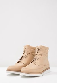 Beżowe skórzane botki z okrągłym noskiem, wyściełanym wsparciem kostki oraz białą gumową podeszwą. Posiadają sznurówki i czysty, minimalistyczny design.
