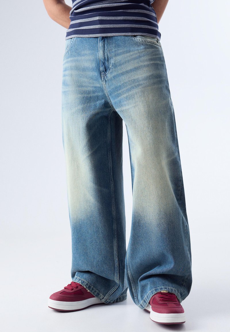 Personne portant un jean bleu délavé à jambes larges, une chemise rayée et des baskets rouges, debout devant un fond clair.