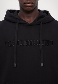 La sudadera negra presenta un logo bordado texturizado "VERSACE JEANS COUTURE" en color a juego, con cordones de contraste y un corte relajado.