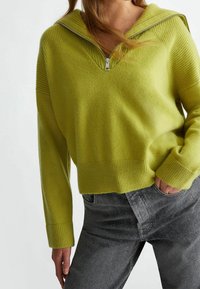 Maglione a maglia verde chiaro con cappuccio e mezza zip, con polsini e orlo a costine; abbinato a jeans grigio scuro.