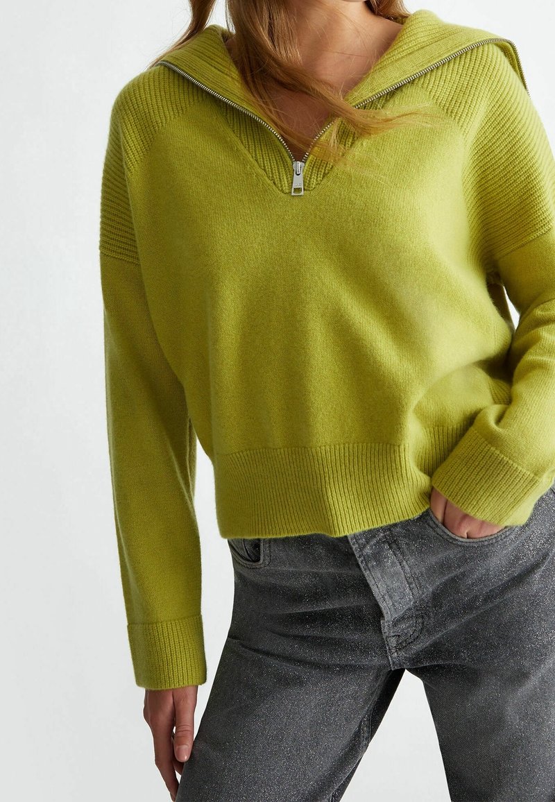 Maglione a maglia verde chiaro con cappuccio e mezza zip, con polsini e orlo a costine; abbinato a jeans grigio scuro.