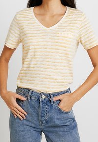 Camiseta de algodón a rayas amarillas y blancas con escote en V, mangas cortas y un bolsillo en el pecho izquierdo, combinada con jeans de tiro alto azules.