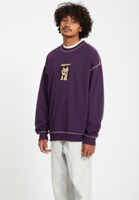 Paarse sweatshirt met een grafische print op de borst, rib gebreide kraag en contrasterende stikseldetails. Gecombineerd met lichtgrijze jeans.