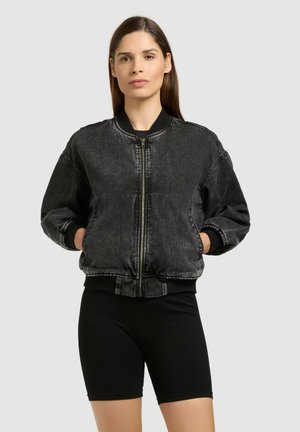 Kvinde med glat brunt hår iført en sort vasket denim bomberjakke og sorte cykelshorts, stående med hænderne i lommerne.