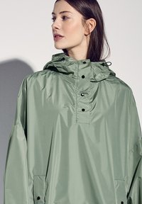 Jasnozielona bluza z wysokim kołnierzem, kapturem, czarnymi zatrzaskami i przednią kieszenią. Gładki, błyszczący materiał o luźnym kroju.