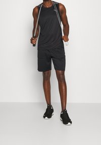 Débardeur de sport noir avec un logo, short noir avec poches, et baskets noires avec des accents blancs, posés avec une corde à sauter.