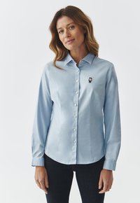 Lichtblauw langemouwen shirt gemaakt van gladde stof, met een kraag en een klein berenembleem op de linkerkant van de borst.