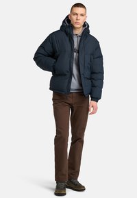 Mørkeblå puffjakke med hætte, med en frontlynlås, elastiske manchetter og en grå sweatshirt under. Brune bukser og grå støvler.