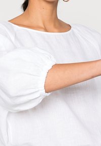 Haut en lin blanc avec un col rond et des manches bouffantes. Le tissu a une apparence douce et texturée avec des poignets élastiques aux manches.