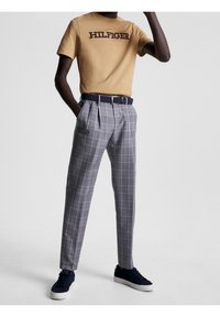 En person iklädd en beige "Hilfiger" t-shirt, grå rutiga byxor med ett svart bälte och svarta sneakers med vita sulor, står mot en vit bakgrund.